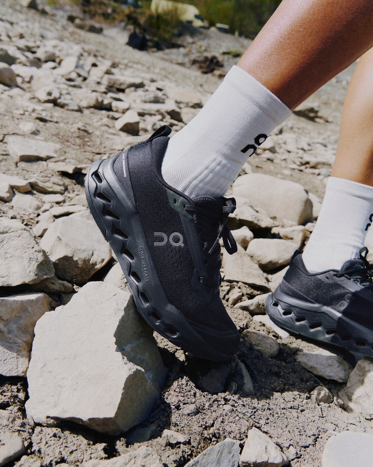 Feet of runner in white socks and black On Cloudsurfer Trail 2 trail-running shoes, running on gray rocks.