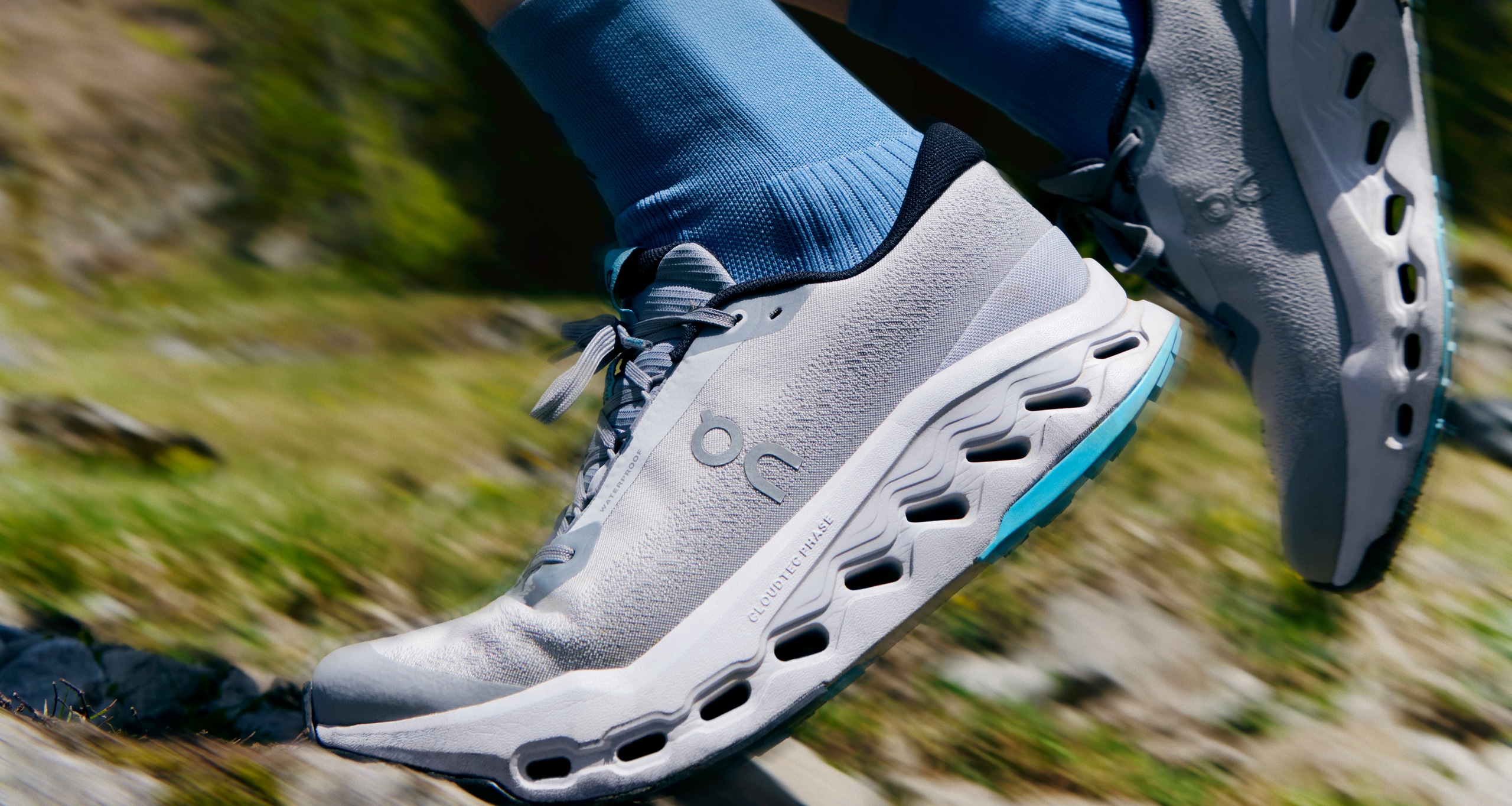 Feet of a runner in blue socks and gray On Cloudsurfer Trail 2 trail-running shoes, running on a green, rocky trail.