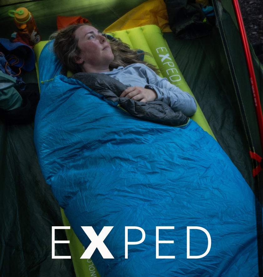 A person in a sleeping bag lays on top of their Exped sleeping pad while looking off into the distance.