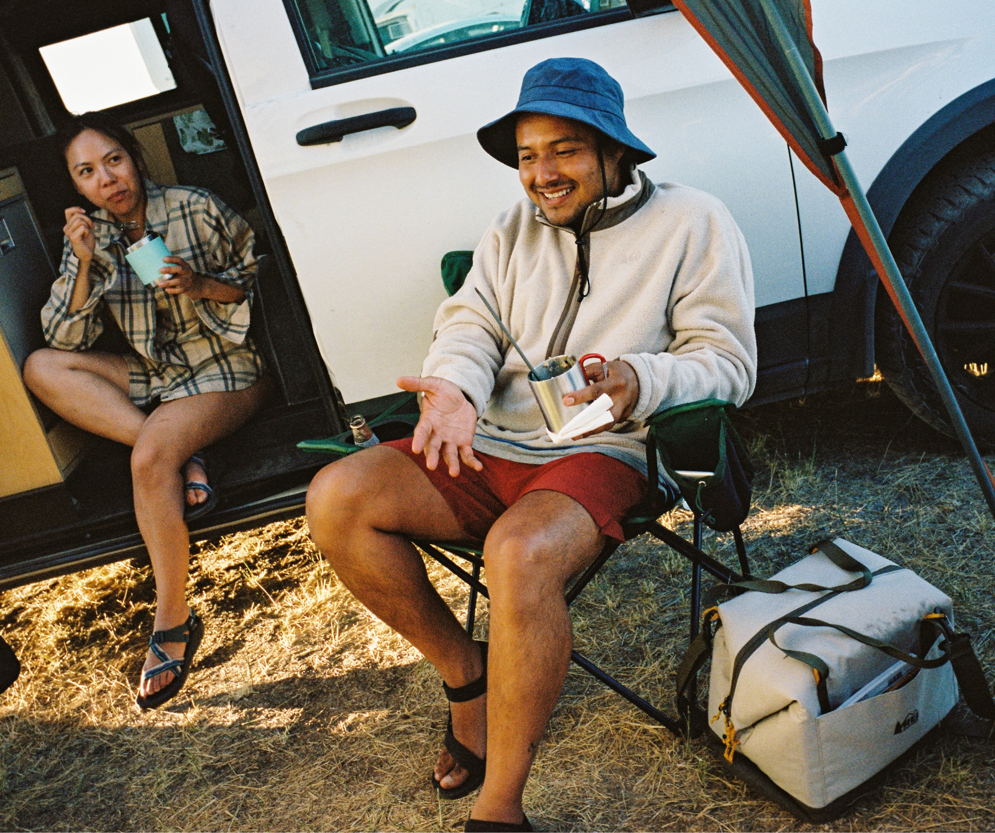 A person sits in a car and enjoys a meal while another person relaxes in a camp chair with a mug.