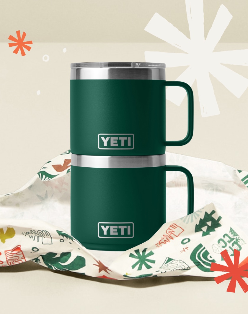 Two stacked YETI mugs sitting on top of torn wrapping paper with holiday motifs.