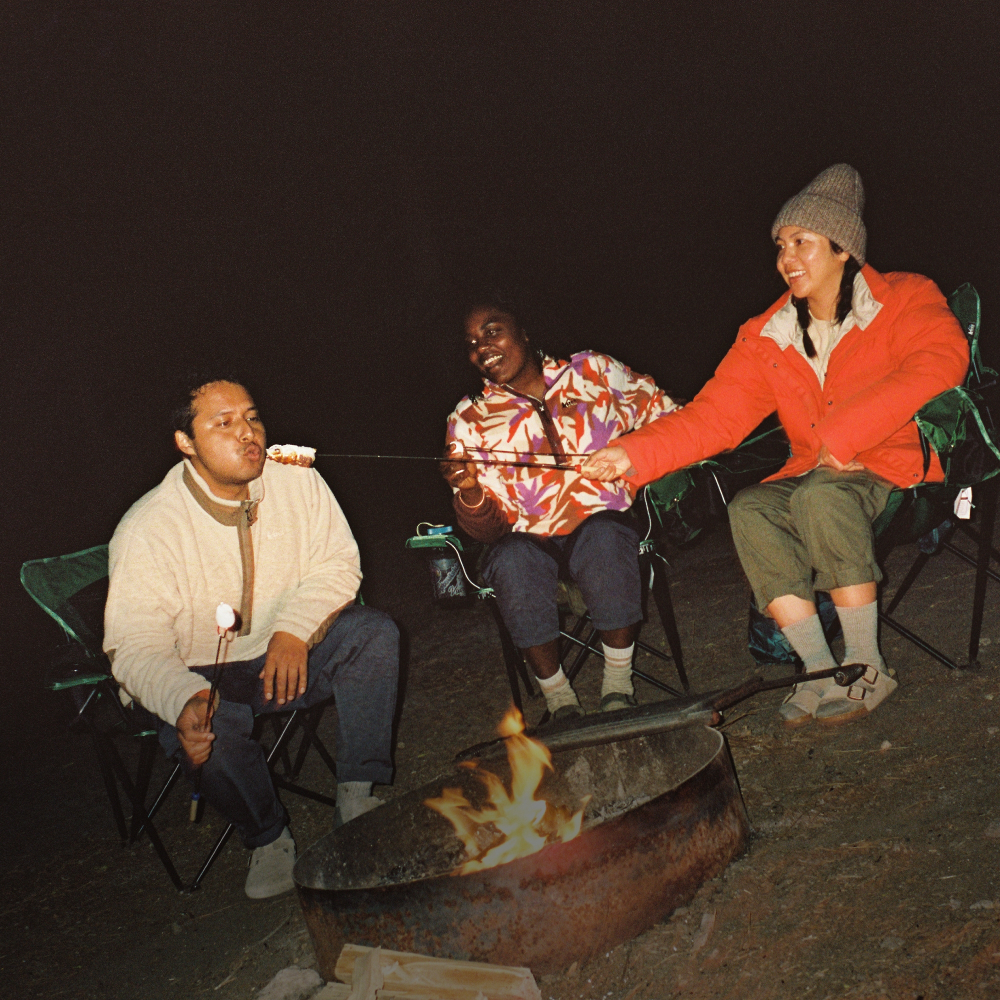 R E I Co-op. Three people gather around a campfire to make s’mores.