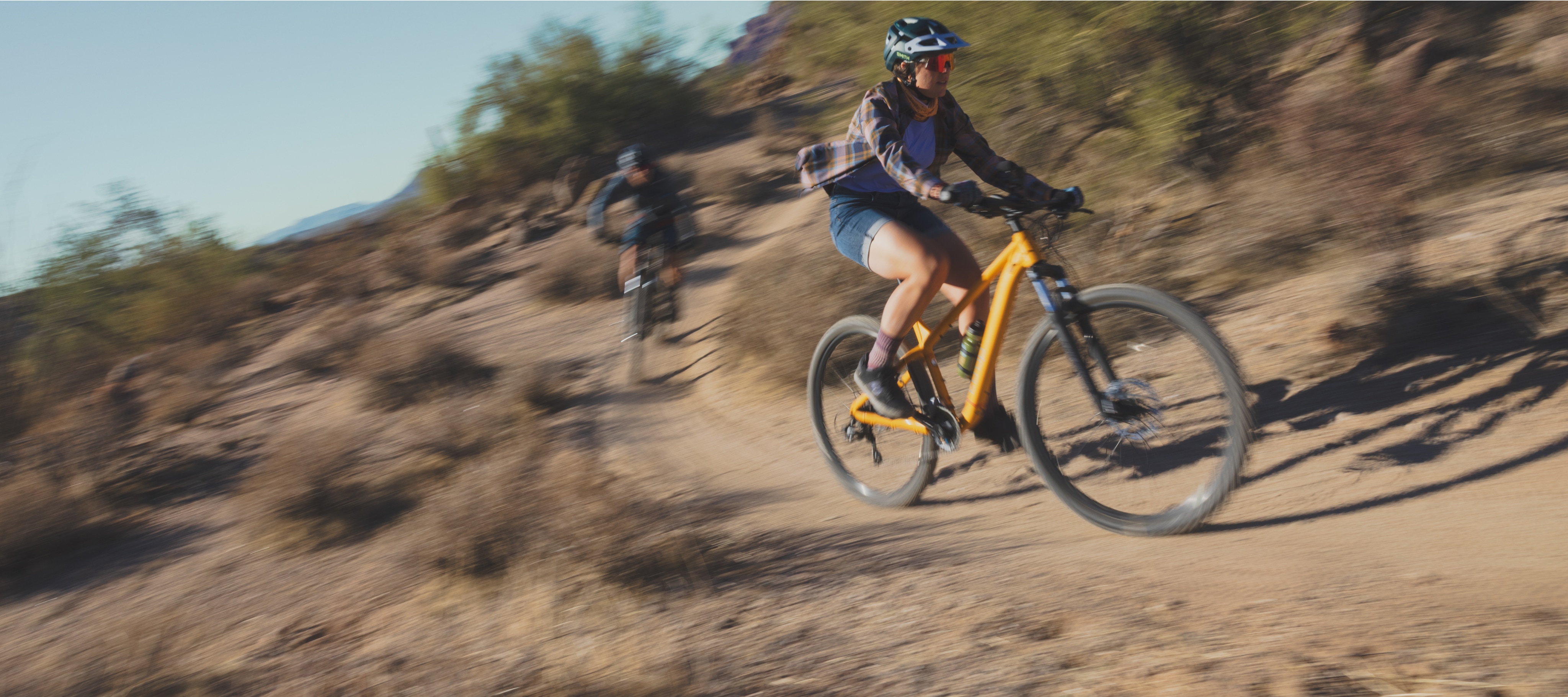 Two people zoom down a desert trail on their bikes.