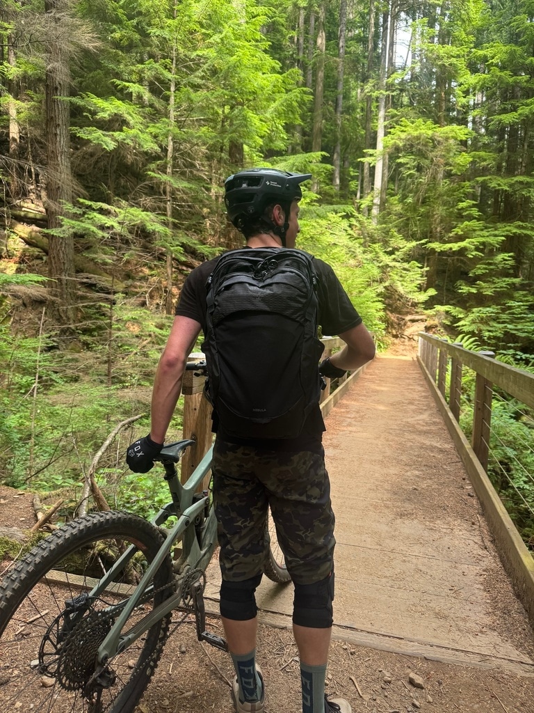 The author stands in front of an on-trail bridge with a mountain bike, wearing the Osprey Nebula backpack.