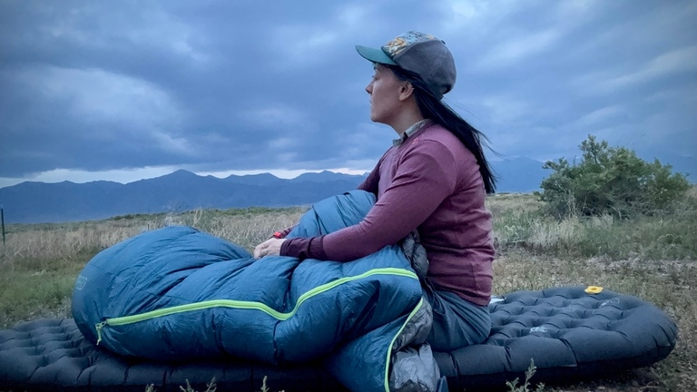 Gear tester and writer, Emily Pennington, sits upright in her Exped Terra 15 sleeping bag. The bag is blue with a green zipper. She's outside and sitting on top of a sleeping pad.