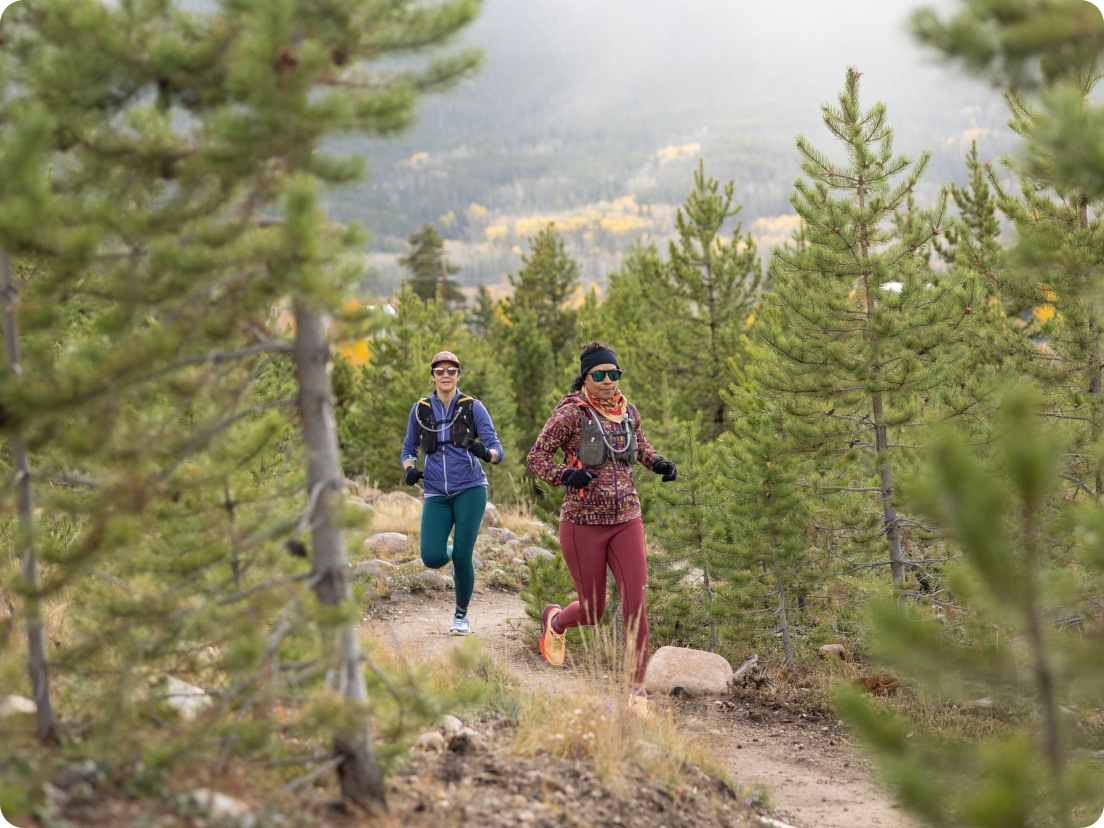 Two people run through a dirt trail lined with trees.