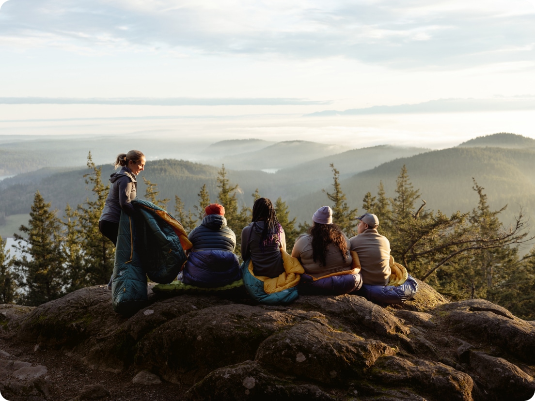 A group of people sit on top of a large rock in their sleeping bags to watch the sun set.