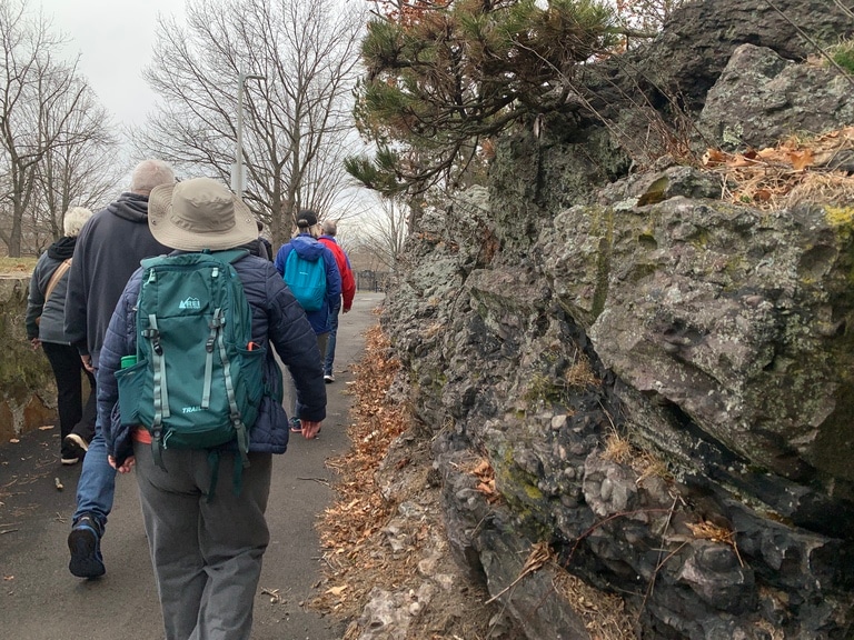 A group of urban hikers pass by a rocky outcropping while walking on a paved trail.
