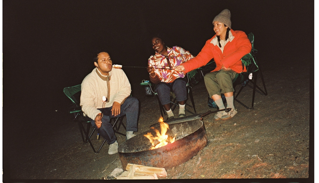 three people eating food around a campfire
