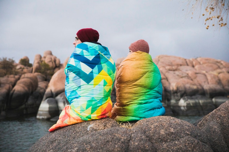 Two people sit together near a body of water. Each of them is wrapped in a colorful Rumpl blanket.