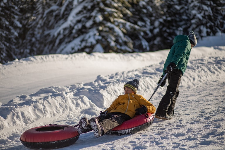 An adult in a green jacket is pulling a child in a snow tube.