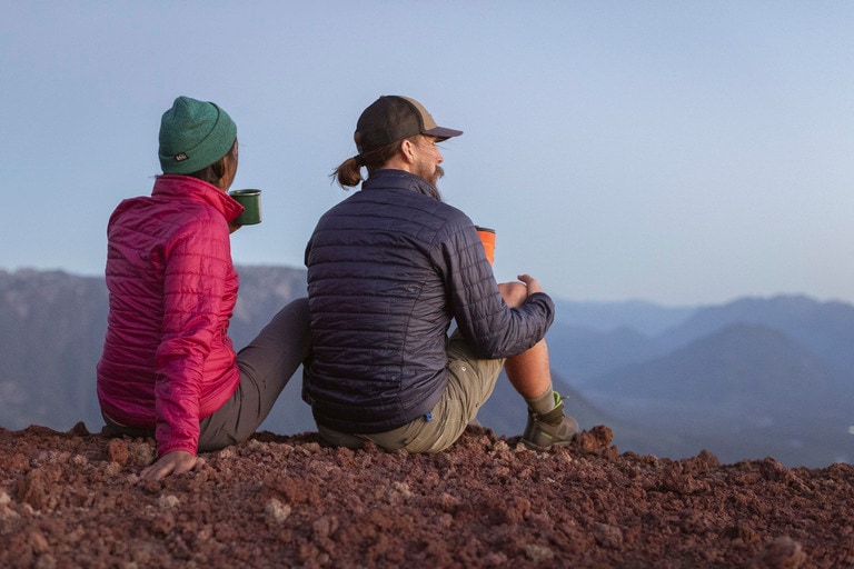 Two backpackers sit on the ground sipping a hot drink in a camp mug.