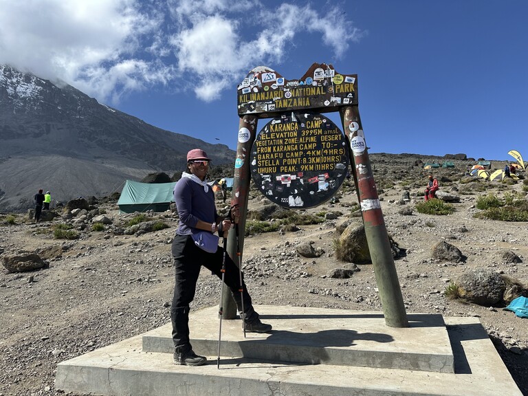 A Black mountaineer poses at a basecamp sign on a mountain.