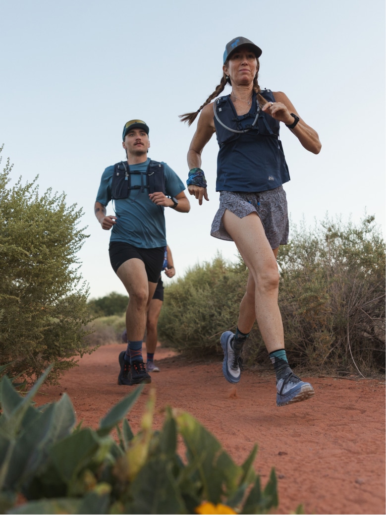 A group of people run through a sandy trail lined with shrubs.