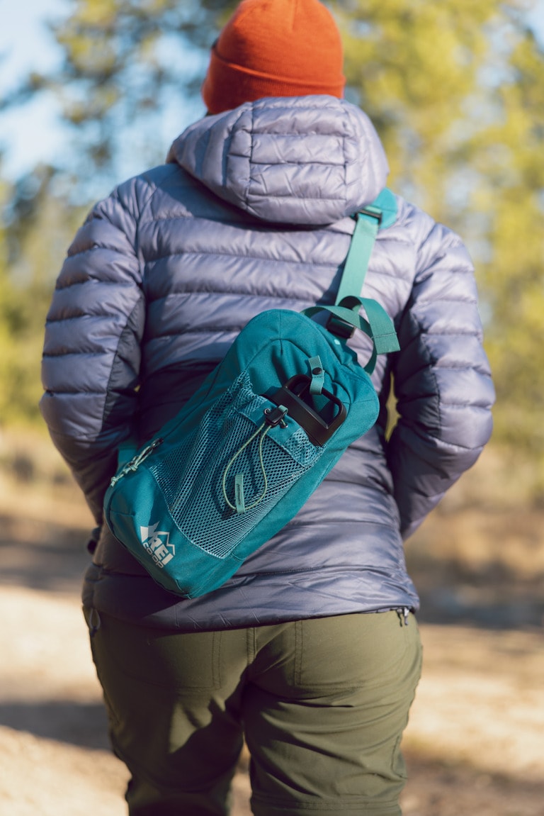 A person wearing a purple puffy jacket walks away from the camera, wearing a blue cross-body bag with a water bottle in it.