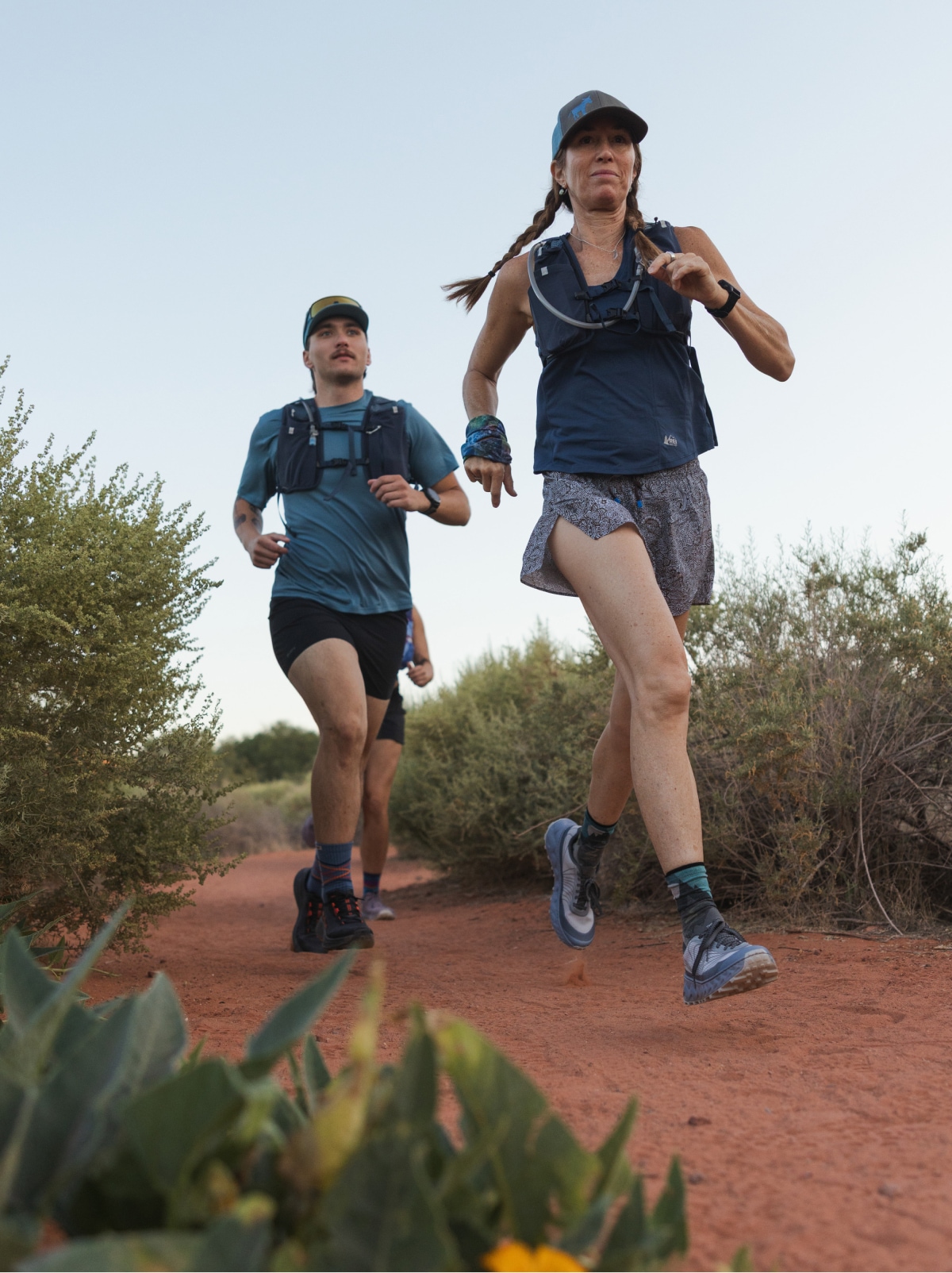 A group of people run through a sandy trail lined with shrubs.