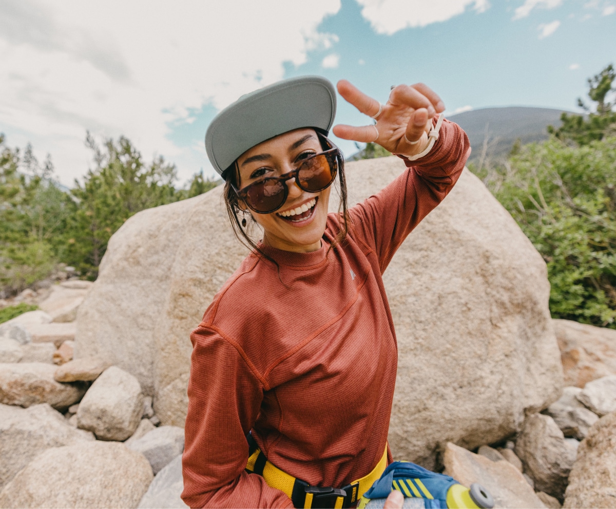 A person throws up a peace sign and smiles for the camera in front of a large rock.