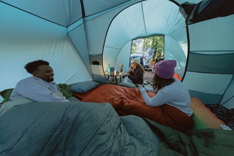 Picture of three campers sitting inside a tent. They are each tucked inside a different sleeping bag.