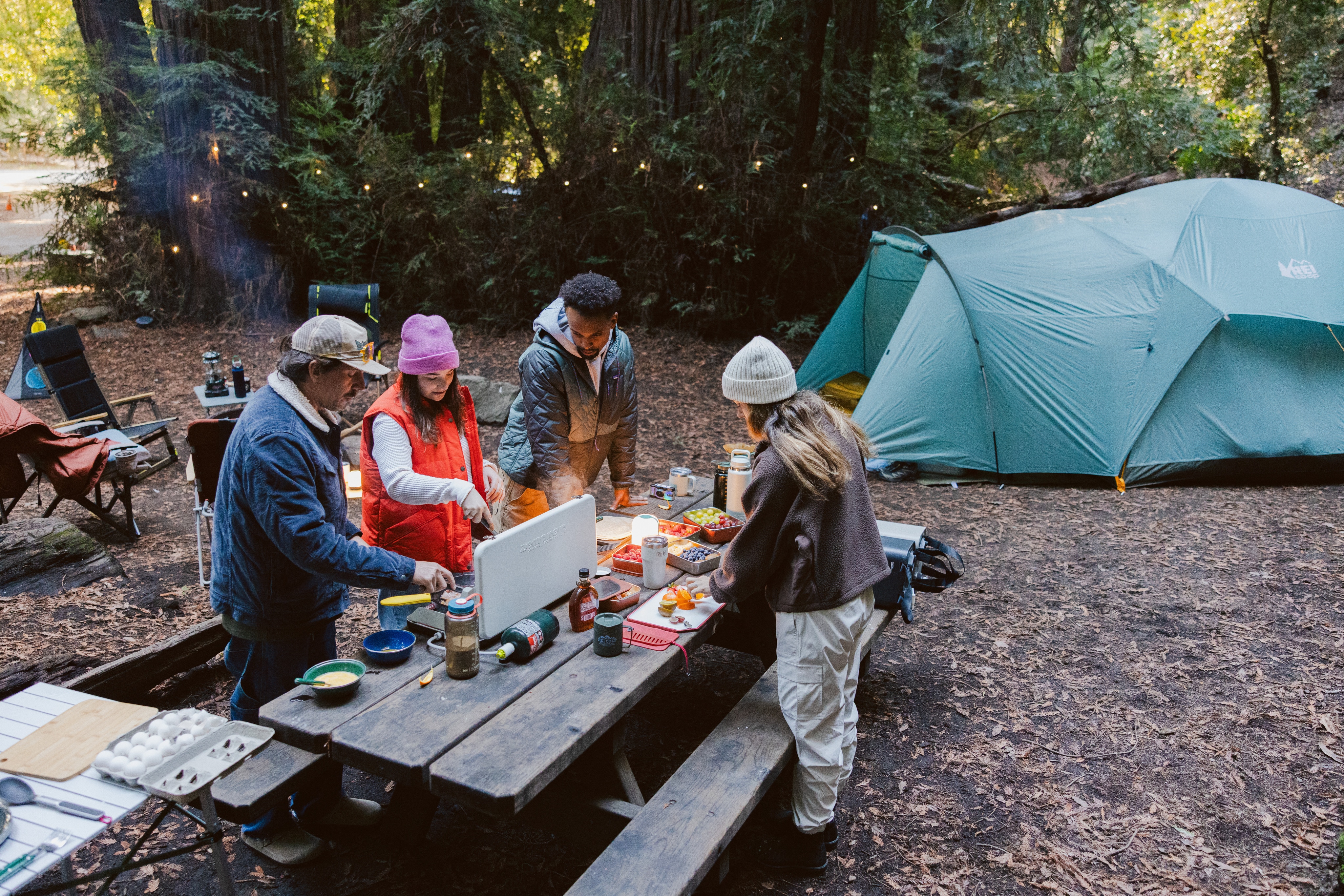 A group of people gather around a table to cook a meal together at their campsite.