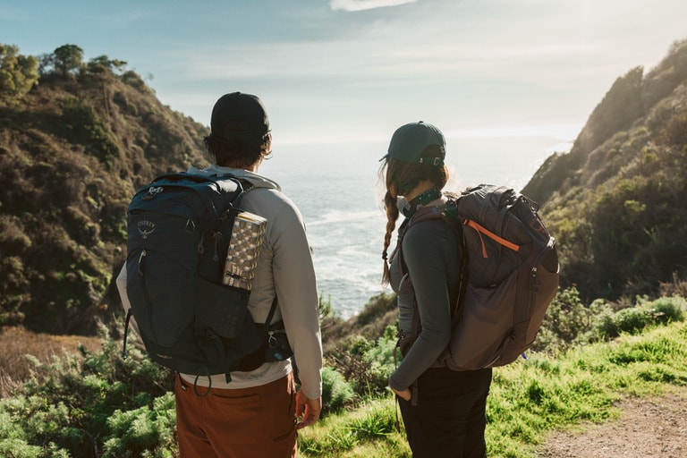 Two people wearing hiking gear and daypacks stand on a cliff overlooking water.