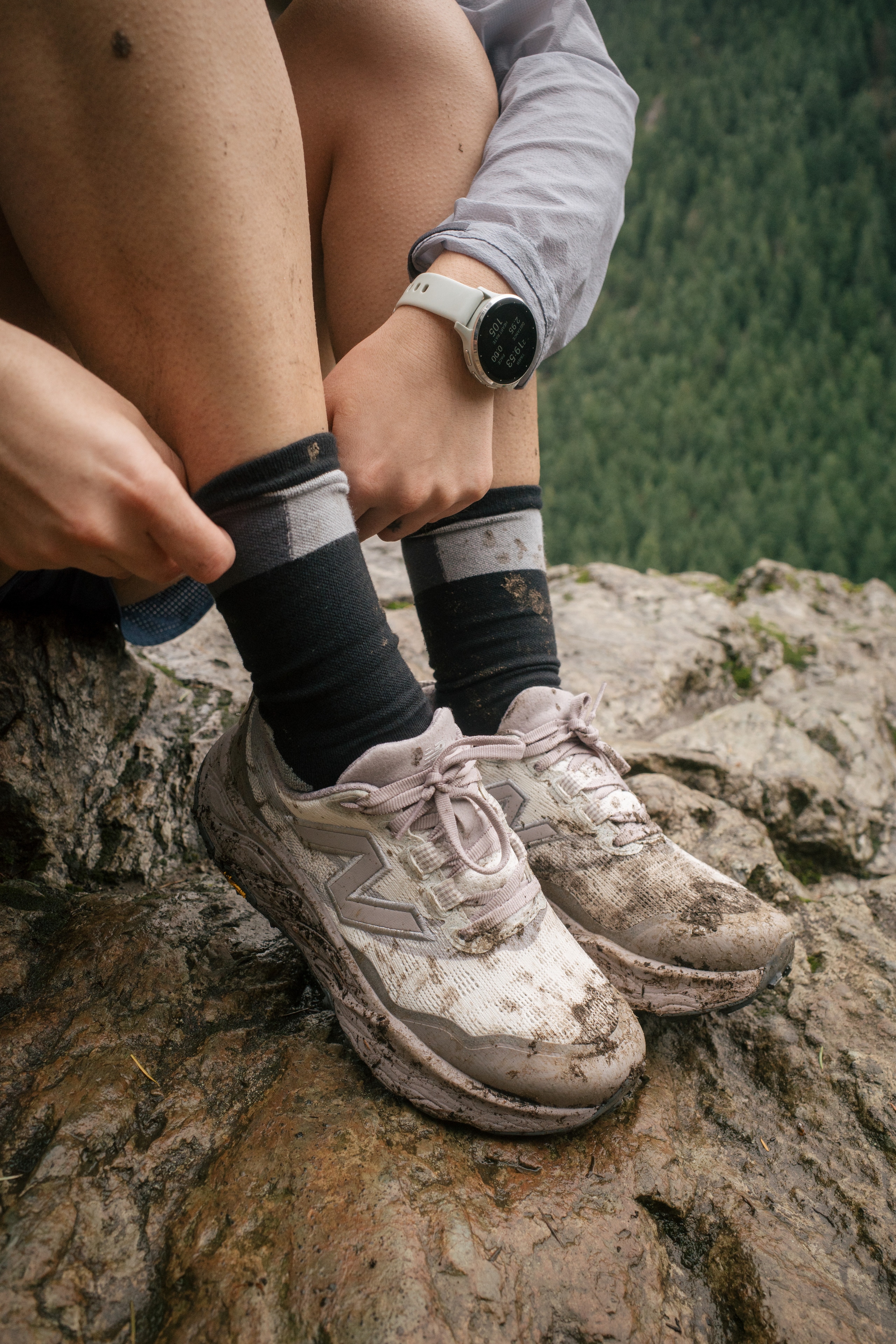 A runner sitting on a muddy rock in a pair of New Balance Fresh Foam X Hierro v9 trail-running shoes in white.