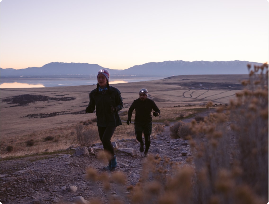 Two people run up a rocky, desert trail while the sun begins to set. 