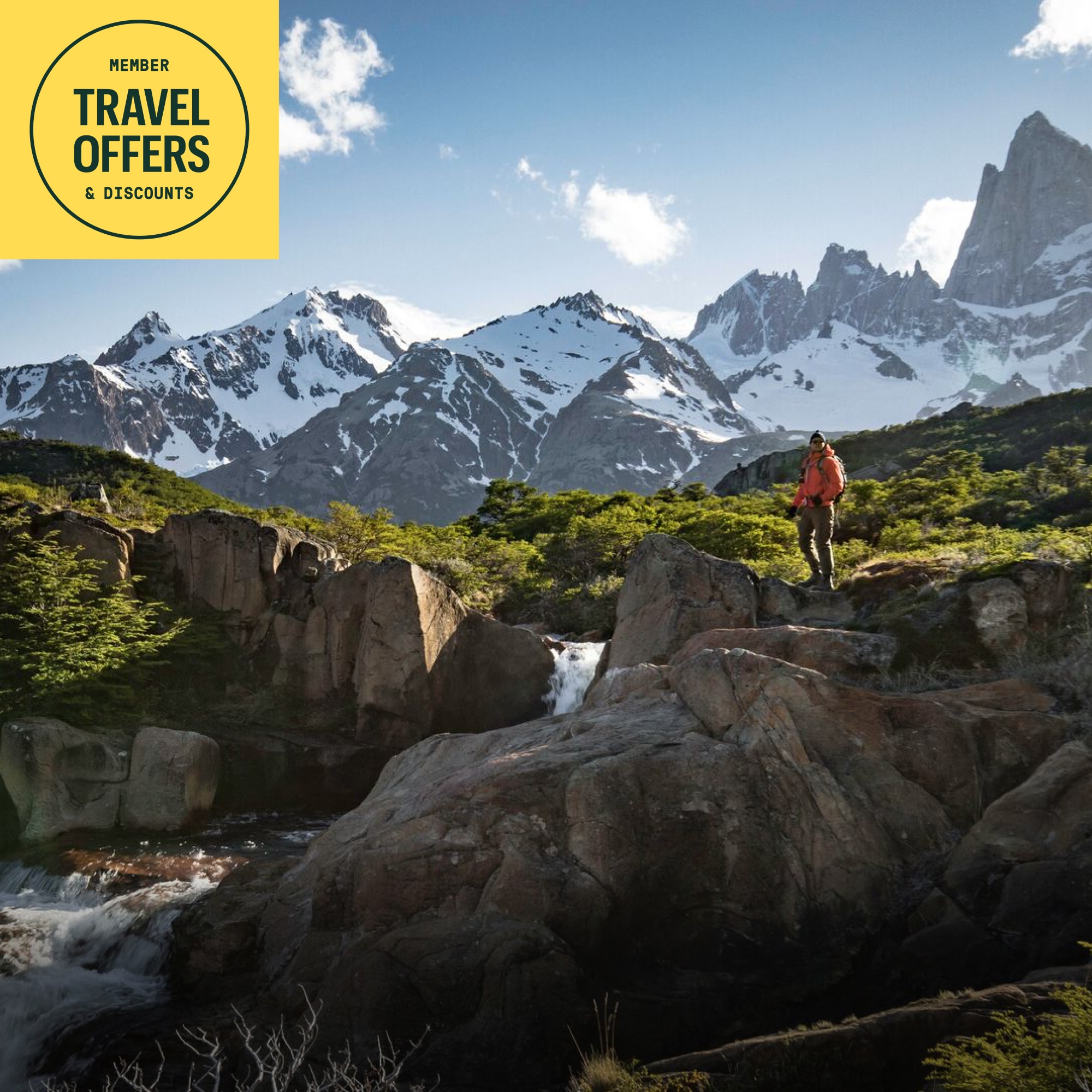 R E I Co-op & Intrepid. A person stands on top of a rock with a snow covered mountain range in the background.