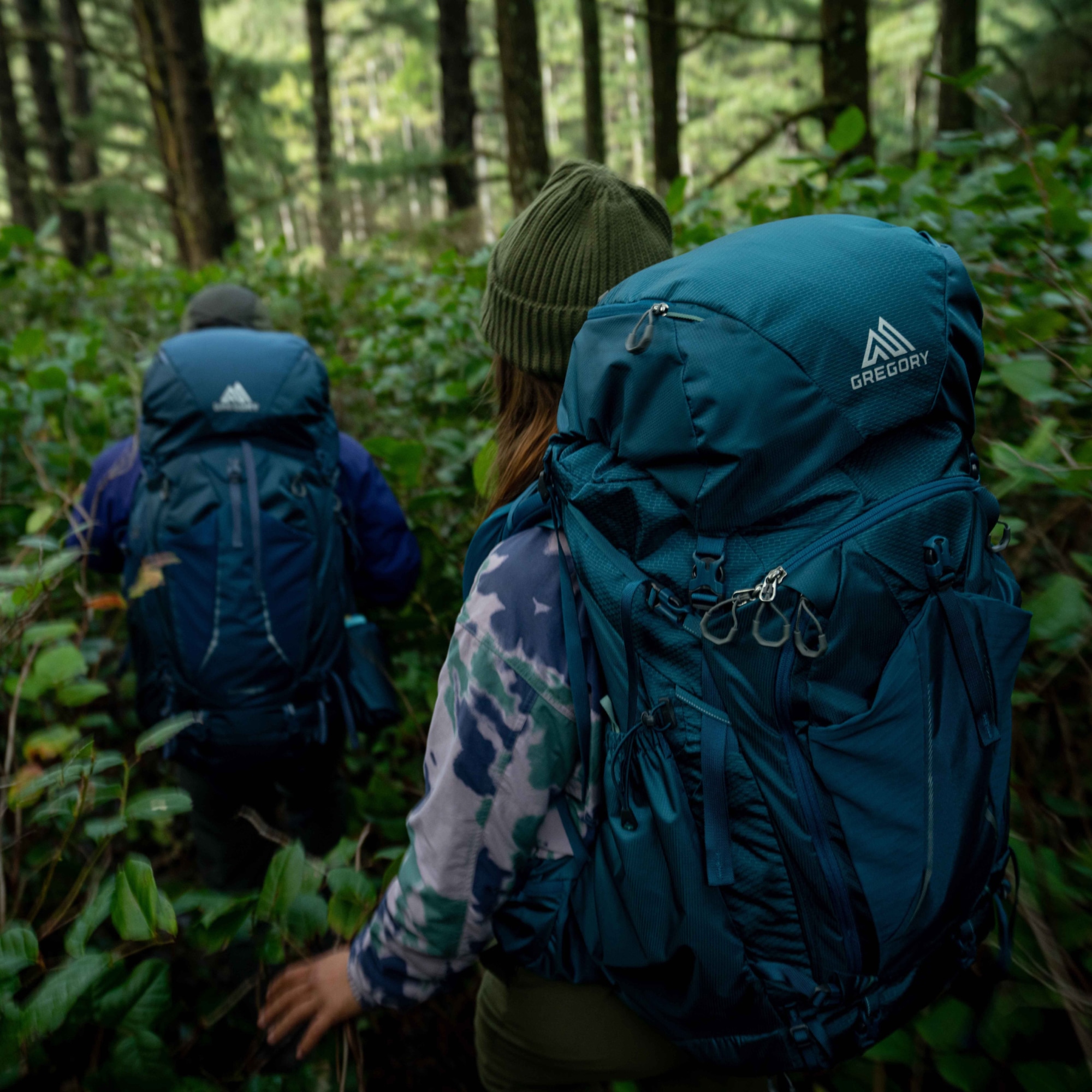 Two people hike through a dense forest with their Gregory packs on.