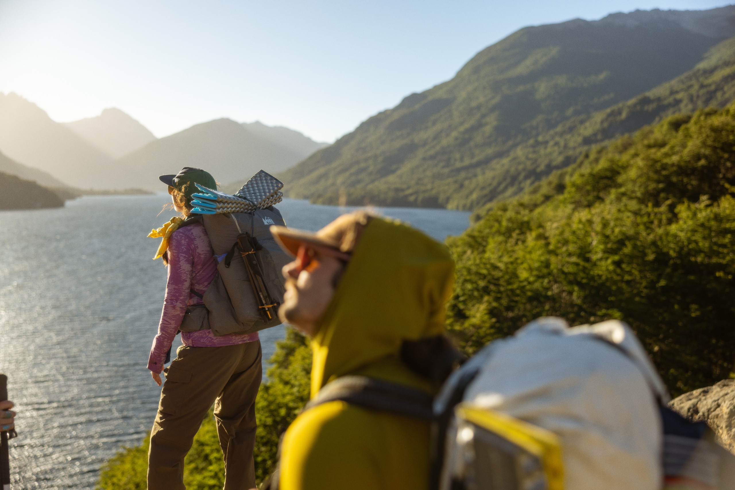 Two people stand on a ridge overlooking a body of water, with the sun shining on their faces.