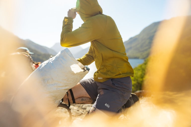 A person kneels down and looks away from the camera while reaching into their top-loading daypack.