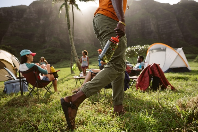 A camper walks past a busy campsite carrying a water bottle.