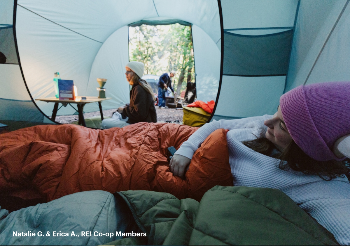A person makes coffee inside of a tent while another person relaxes in their sleeping bag.