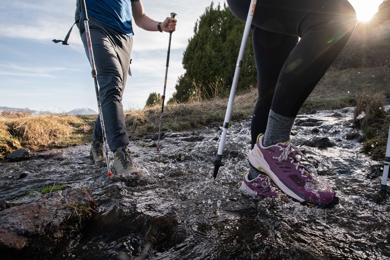 Two hikers cross water in low-cut hiking shoes at sunset