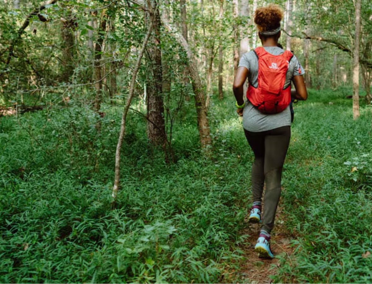 A person wearing a hydration pack runs through a trail in a forest.