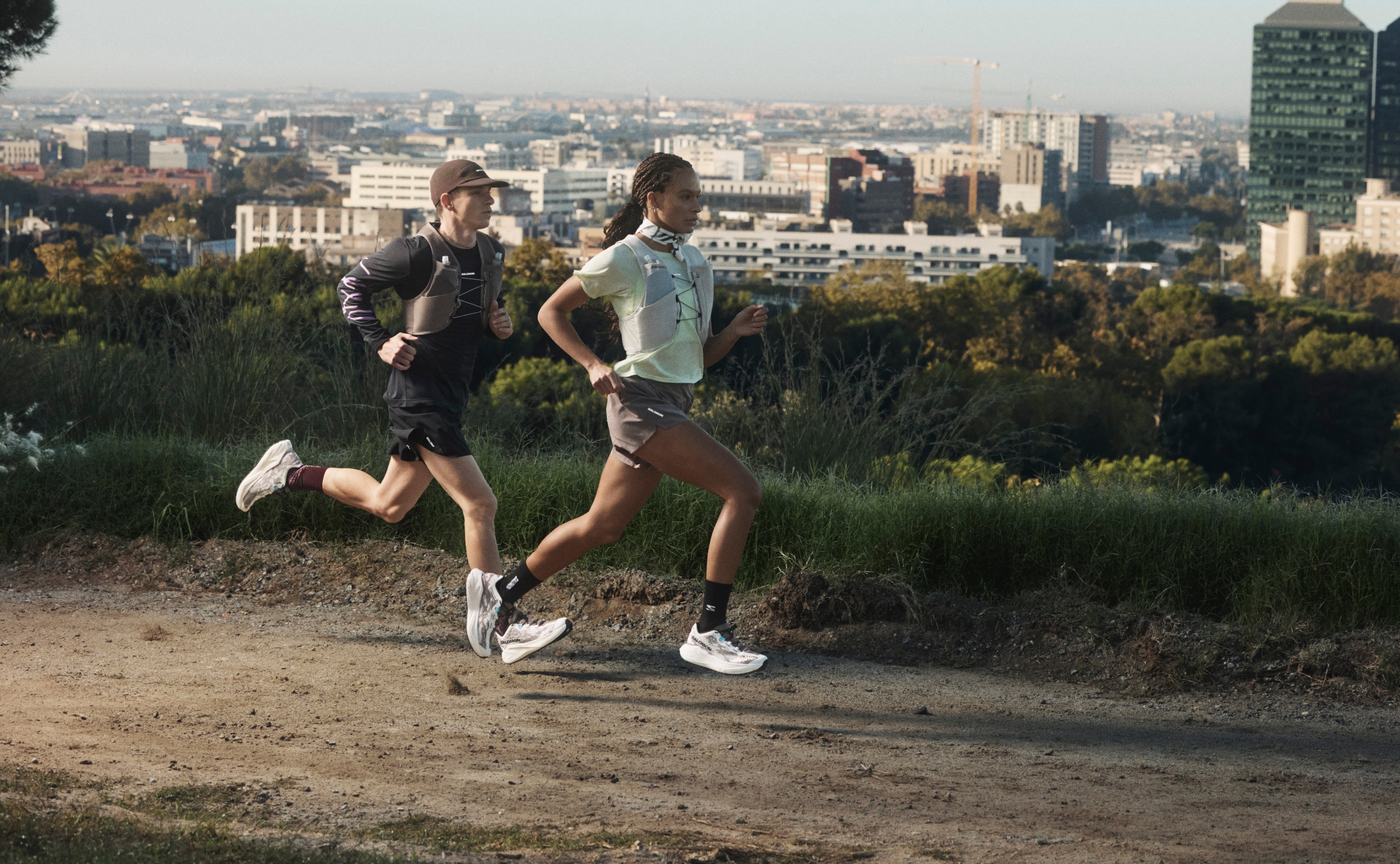 Two people run on a dirt trail with a view of a city behind them.