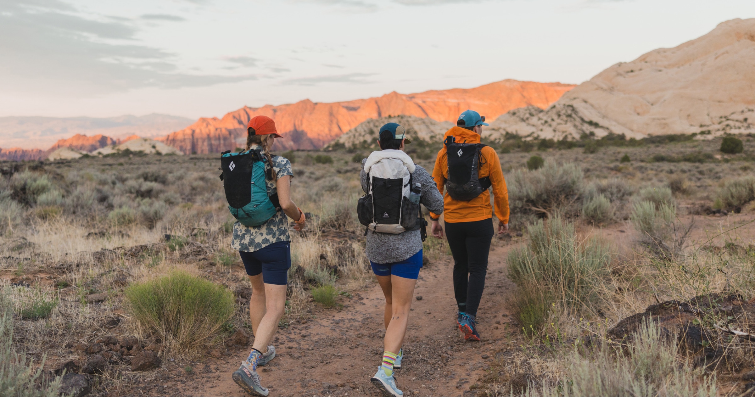 Three people walk through a desert trail while the sun begins to set.