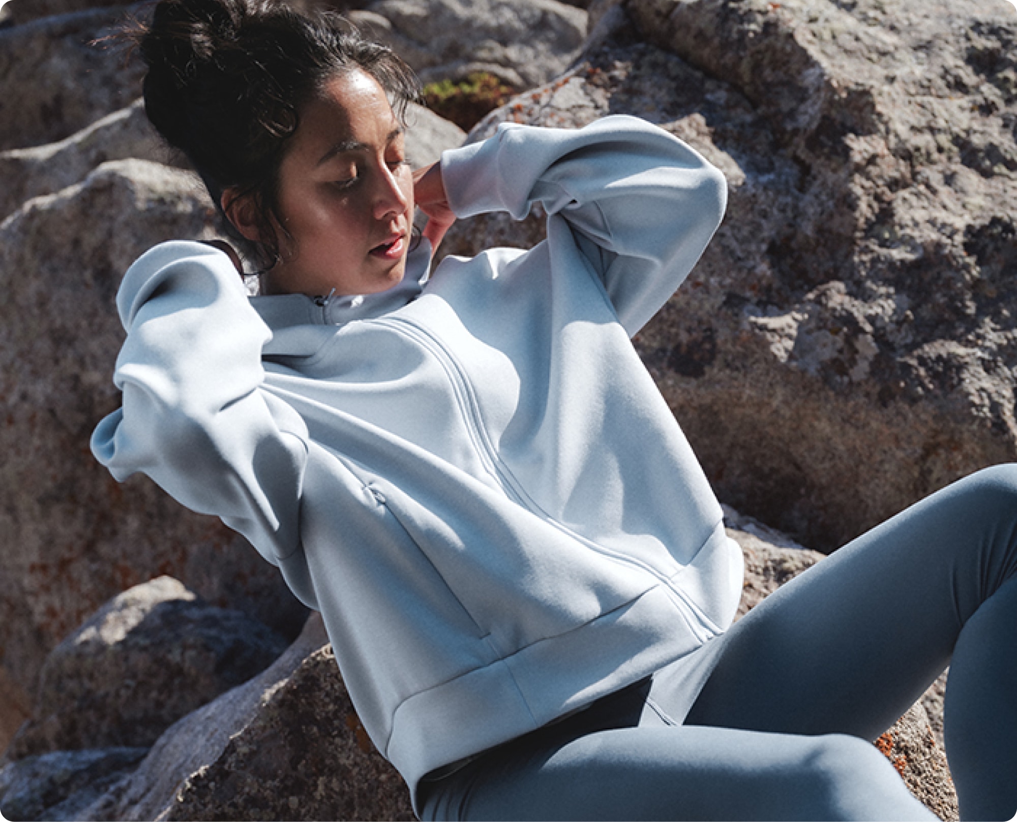 A person adjusts the neckline on their jacket while sitting on a large rock.