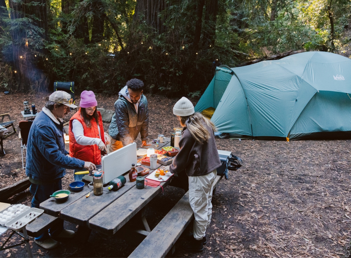 A group of people gather around a table and cook a meal together at their campsite.
