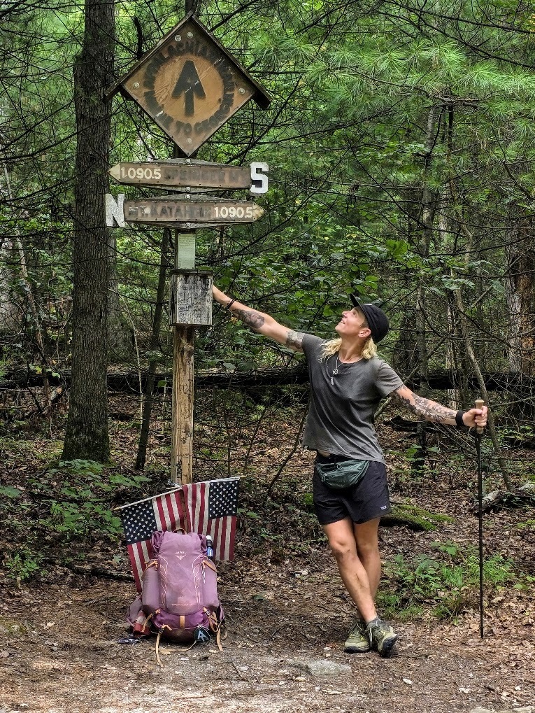 A person stands at a trail marker along the Appalachian Trail. Her purple pack is at the base and she holds a trekking pole in one hand.