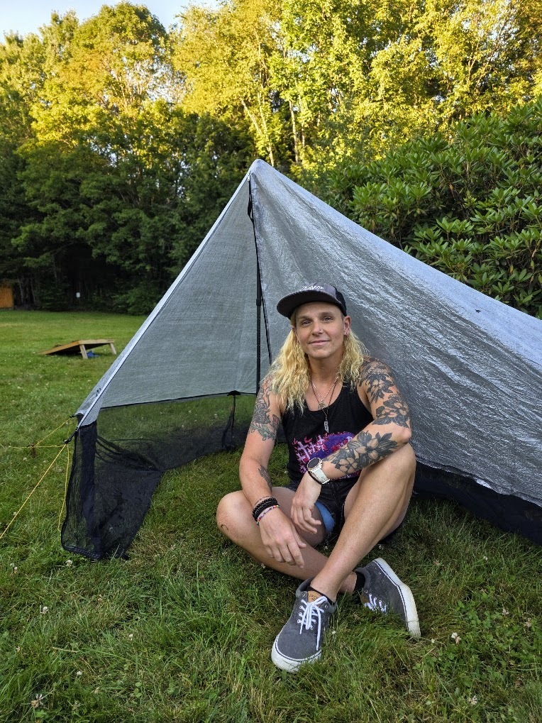 A person with blonde hair and tattoos sits underneath a lightweight tarp shelter on a green space in front of trees.
