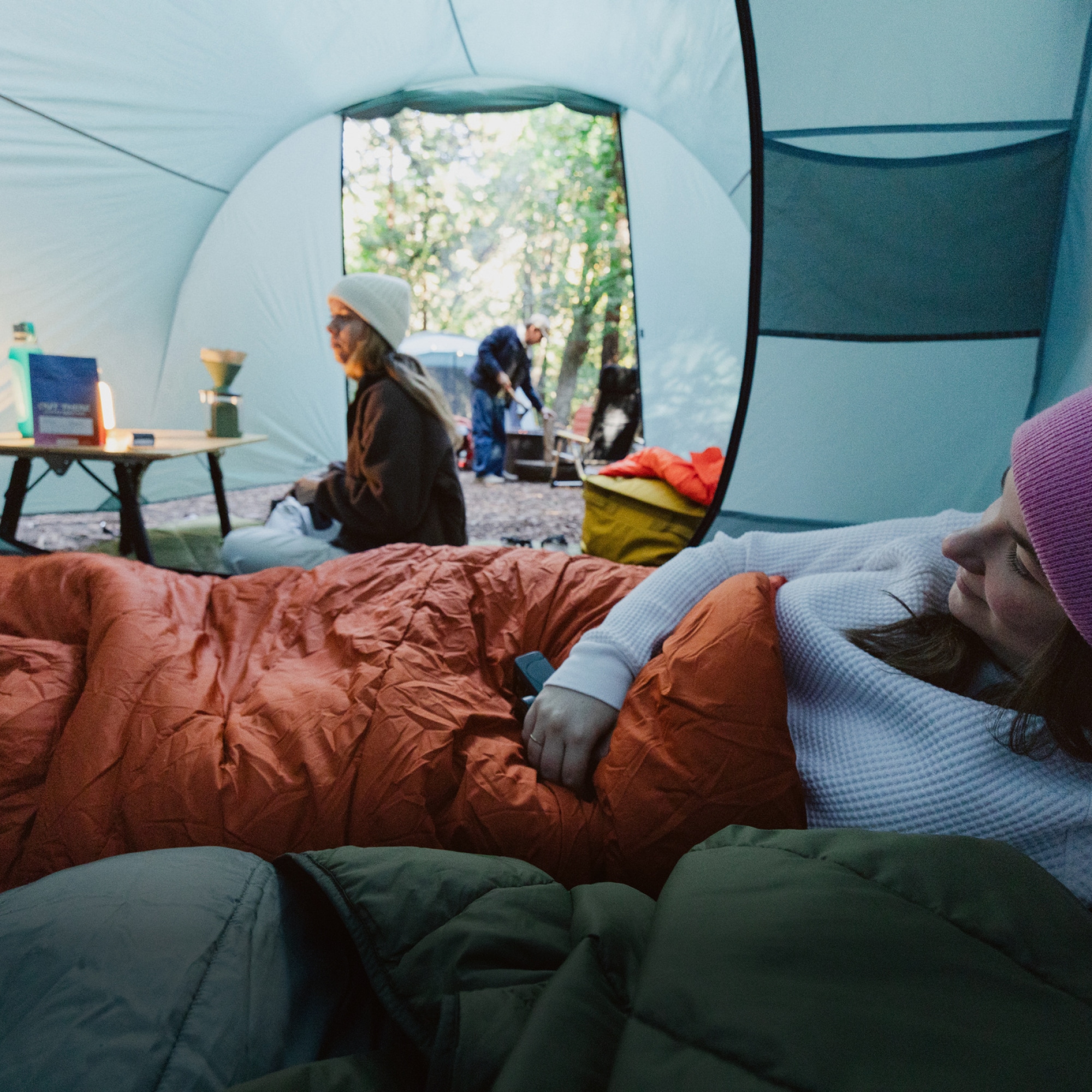Two people inside of a tent. One relaxes in a sleeping bag while the other makes coffee.