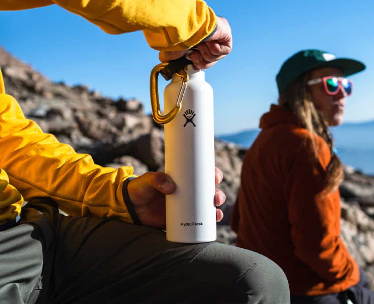 A person sits down while opening up their Hydro Flask water bottle while another person looks off into the distance in the background.