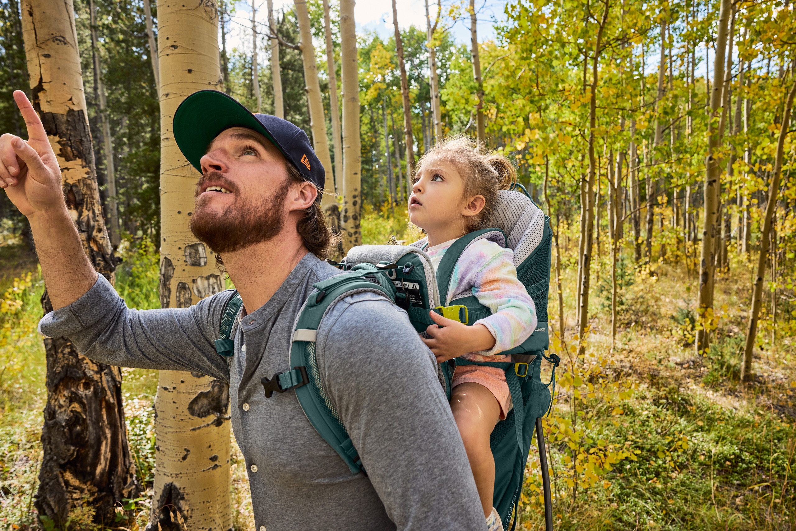 A person carries a child in a carrier backpack through a wooded area