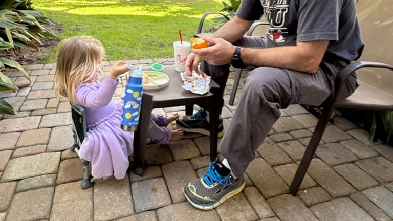 A little girl sits in a small chair and holds a blue rainbow water bottle next to an adult.