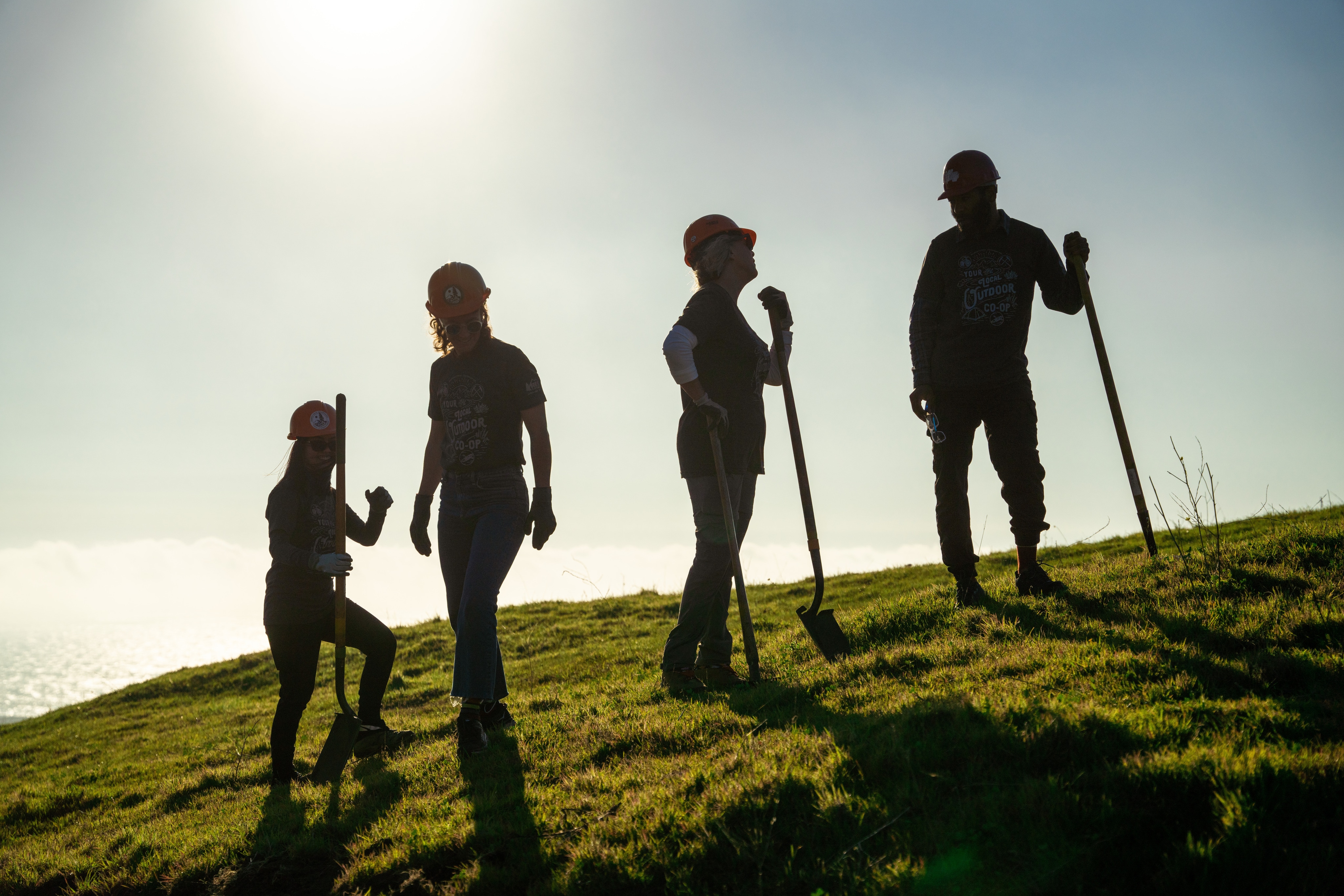 Four people with shovels stand on a grassy hillside, silhouetted by the sun.