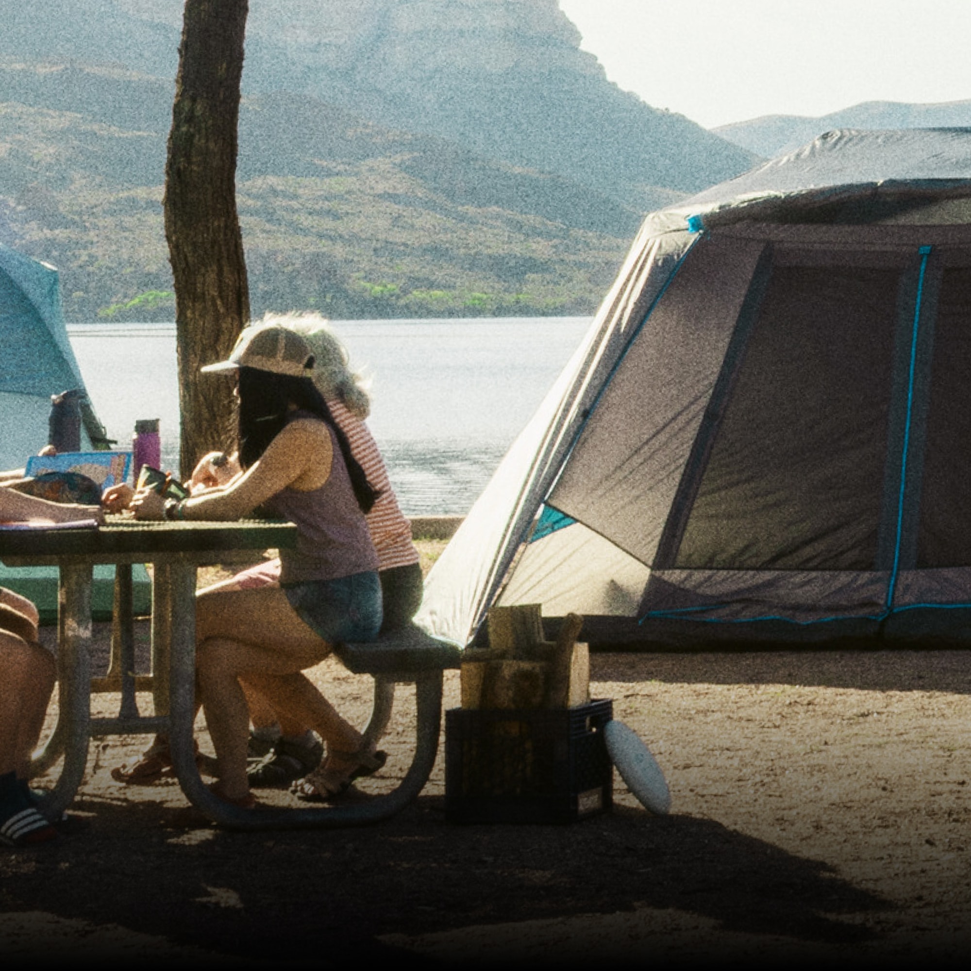 R E I Co-op & CORE Equipment. Two people sit on one side of a table with a tent in the background.