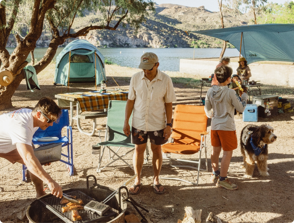 A person grills meat at their campsite while someone watches with others gather around a table in the background.
