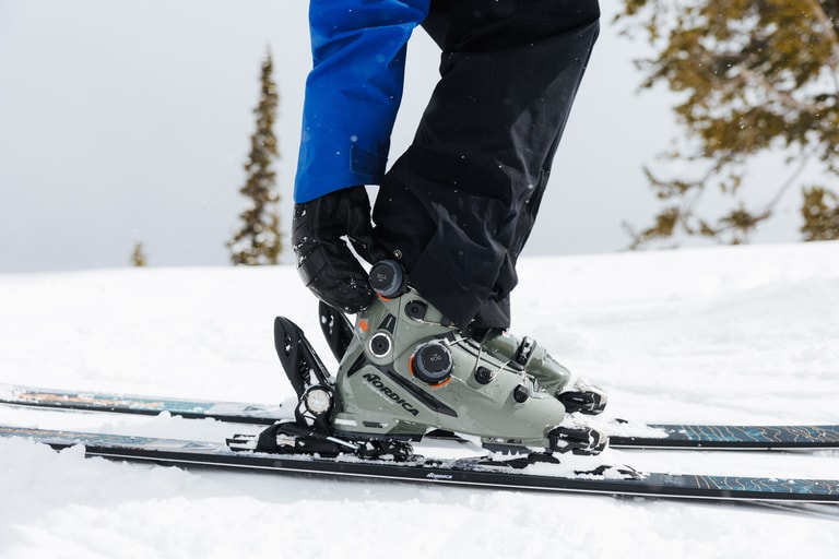 A skier reaches down to adjust the dials on their downhill ski boots.