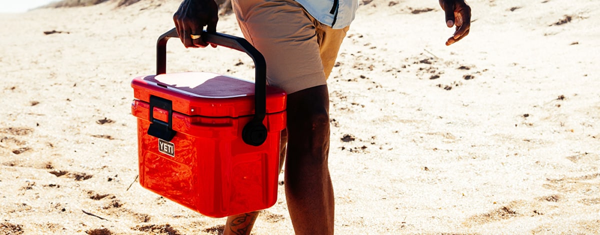 A person walks along a sandy path while carrying a red YETI Roadie 8 cooler.