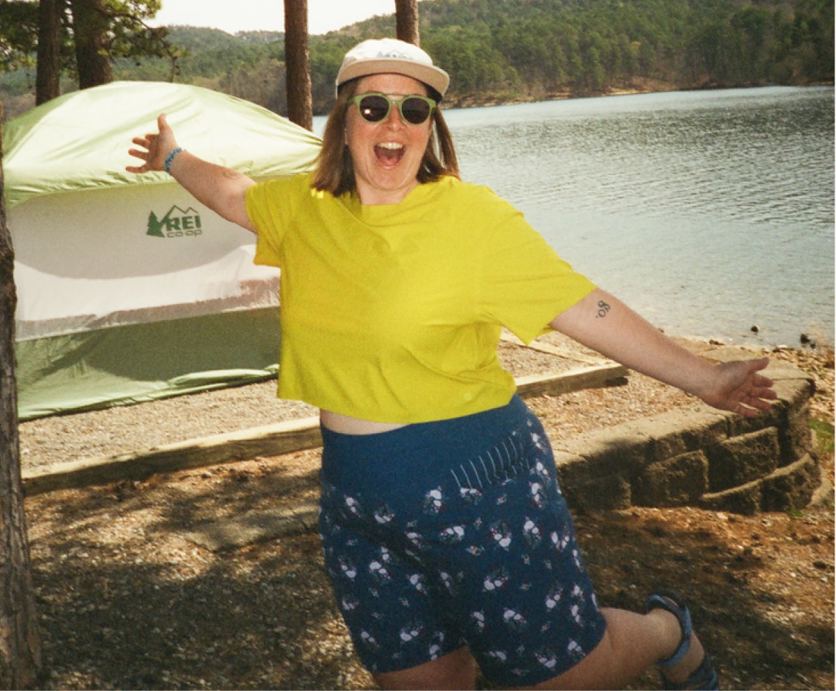 A person smiles for the camera in front of a green tent with a lake in the background.